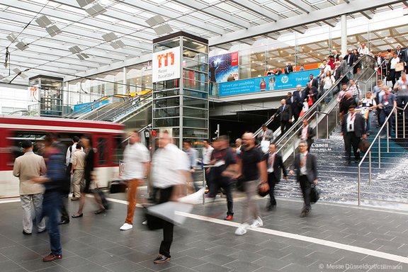 Menschen strömen die Treppen und Rolltreppen der Messe Düsseldorf hinauf und hinunter, dynamisches Messeumfeld.